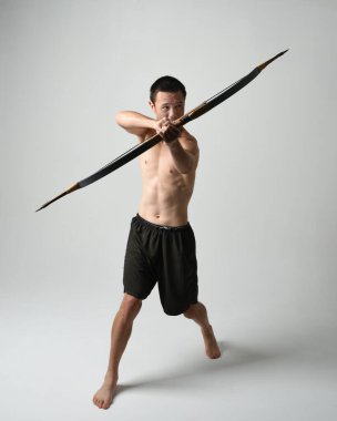 Asyalı erkek modelin tam boy portresi, av yayı ve okçuluk silahı taşıyor, savaşçı eğitim pozisyonunda duruyor, beyaz stüdyo arka planında izole edilmiş..