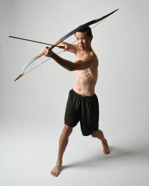 Asyalı erkek modelin tam boy portresi, av yayı ve okçuluk silahı taşıyor, savaşçı eğitim pozisyonunda duruyor, beyaz stüdyo arka planında izole edilmiş..