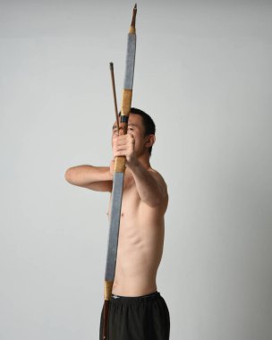 Asyalı erkek modelin tam boy portresi, av yayı ve okçuluk silahı taşıyor, savaşçı eğitim pozisyonunda duruyor, beyaz stüdyo arka planında izole edilmiş..