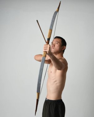 Asyalı erkek modelin tam boy portresi, av yayı ve okçuluk silahı taşıyor, savaşçı eğitim pozisyonunda duruyor, beyaz stüdyo arka planında izole edilmiş..