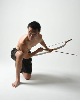 Asyalı erkek modelin tam boy portresi, av yayı ve okçuluk silahı taşıyor, savaşçı eğitim pozisyonunda duruyor, beyaz stüdyo arka planında izole edilmiş..
