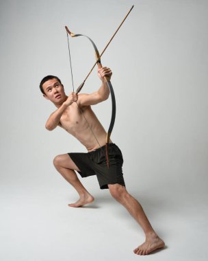 Asyalı erkek modelin tam boy portresi, av yayı ve okçuluk silahı taşıyor, savaşçı eğitim pozisyonunda duruyor, beyaz stüdyo arka planında izole edilmiş..