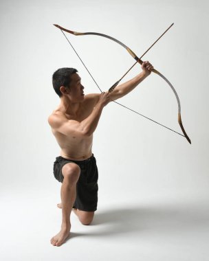Asyalı erkek modelin tam boy portresi, av yayı ve okçuluk silahı taşıyor, savaşçı eğitim pozisyonunda duruyor, beyaz stüdyo arka planında izole edilmiş..