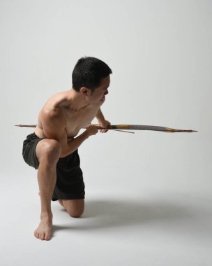 Asyalı erkek modelin tam boy portresi, av yayı ve okçuluk silahı taşıyor, savaşçı eğitim pozisyonunda duruyor, beyaz stüdyo arka planında izole edilmiş..