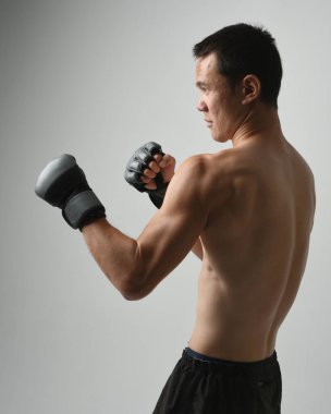 Formda Asyalı erkek modelin yakın portresi, kaslı üstsüz. Spor stenografisi boks eldiveni takıyor, hareketli yumruk pozu veriyor. Beyaz bir stüdyoda izole edilmiş..