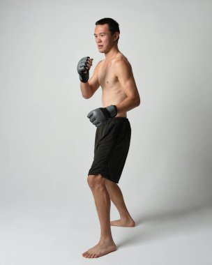Formda, Asyalı bir erkek modelin tam boy portresi, kaslı üstsüz. Spor stenografisi boks eldiveni takıyor, hareketli yumruk pozu veriyor. Beyaz bir stüdyoda izole edilmiş..