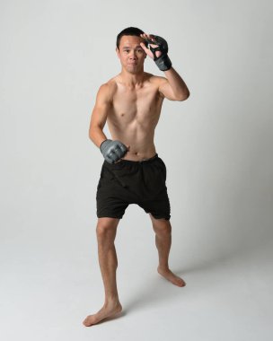 Formda, Asyalı bir erkek modelin tam boy portresi, kaslı üstsüz. Spor stenografisi boks eldiveni takıyor, hareketli yumruk pozu veriyor. Beyaz bir stüdyoda izole edilmiş..