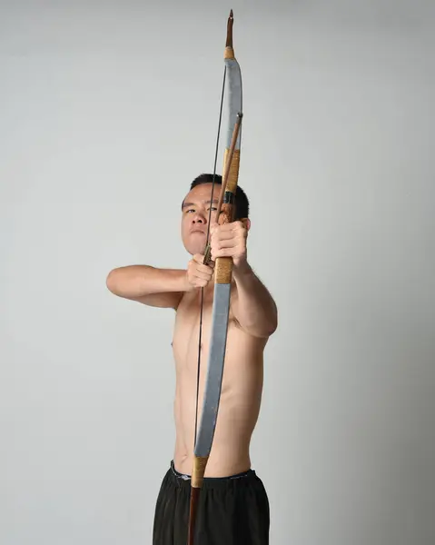 Full length portrait of fit  asian male model,  Holding hunting bow and arrow archery weapon, standing in warrior training action pose, isolated on white studio background.