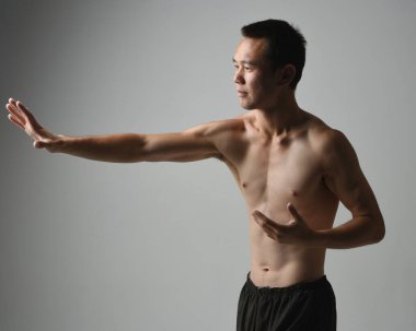 Formda Asyalı erkek modelin yakın portresi, kaslı üstsüz. Jest ti chi kollarını uzatarak poz veriyor. Beyaz bir stüdyonun arka planında karamsar bir siluetle izole edilmiş..