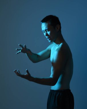 Formda Asyalı erkek modelin yakın portresi, kaslı üstsüz. Jest ti chi 'den esinlenilmiştir. Kollarını uzatarak sanki sihirli bir büyü yapıyor gibi. Huysuz, koyu yeşil bir stüdyonun arka planında, siluetle izole edilmiş..