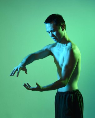 Formda Asyalı erkek modelin yakın portresi, kaslı üstsüz. Jest ti chi 'den esinlenilmiştir. Kollarını uzatarak sanki sihirli bir büyü yapıyor gibi. Huysuz, koyu yeşil bir stüdyonun arka planında, siluetle izole edilmiş..