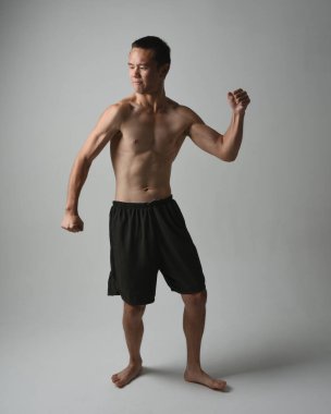 Formda, Asyalı bir erkek modelin tam boy portresi, kaslı üstsüz. Jest ti chi kollarını uzatarak poz veriyor. Beyaz bir stüdyonun arka planında karamsar bir siluetle izole edilmiş..