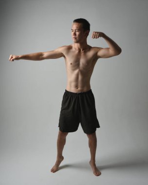 Formda, Asyalı bir erkek modelin tam boy portresi, kaslı üstsüz. Jest ti chi kollarını uzatarak poz veriyor. Beyaz bir stüdyonun arka planında karamsar bir siluetle izole edilmiş..