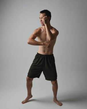 Formda, Asyalı bir erkek modelin tam boy portresi, kaslı üstsüz. Jest ti chi kollarını uzatarak poz veriyor. Beyaz bir stüdyonun arka planında karamsar bir siluetle izole edilmiş..