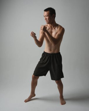 Formda, Asyalı bir erkek modelin tam boy portresi, kaslı üstsüz. Jest ti chi kollarını uzatarak poz veriyor. Beyaz bir stüdyonun arka planında karamsar bir siluetle izole edilmiş..