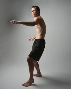 Formda, Asyalı bir erkek modelin tam boy portresi, kaslı üstsüz. Jest ti chi kollarını uzatarak poz veriyor. Beyaz bir stüdyonun arka planında karamsar bir siluetle izole edilmiş..