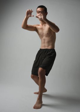 Formda, Asyalı bir erkek modelin tam boy portresi, kaslı üstsüz. Jest ti chi kollarını uzatarak poz veriyor. Beyaz bir stüdyonun arka planında karamsar bir siluetle izole edilmiş..