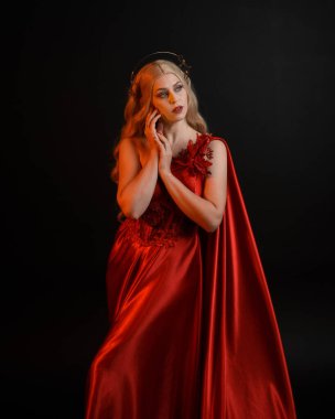 Kırmızı ipek toga elbisesi ve taç giyen, antik mitolojik fantezi tanrıçası gibi giyinmiş güzel sarışın mankenin yakın portresi. Karanlık stüdyo arka planında izole zarif bir poz..