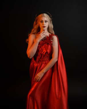 Kırmızı ipek toga elbisesi ve taç giyen, antik mitolojik fantezi tanrıçası gibi giyinmiş güzel sarışın mankenin yakın portresi. Karanlık stüdyo arka planında izole zarif bir poz..