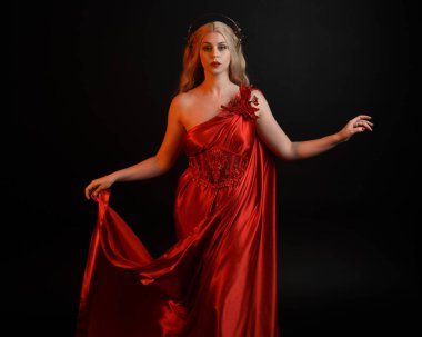 Kırmızı ipek toga elbisesi ve taç giyen, antik mitolojik fantezi tanrıçası gibi giyinmiş güzel sarışın mankenin yakın portresi. Karanlık stüdyo arka planında izole zarif bir poz..