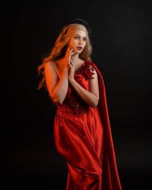 Kırmızı ipek toga elbisesi ve taç giyen, antik mitolojik fantezi tanrıçası gibi giyinmiş güzel sarışın mankenin yakın portresi. Karanlık stüdyo arka planında izole zarif bir poz..