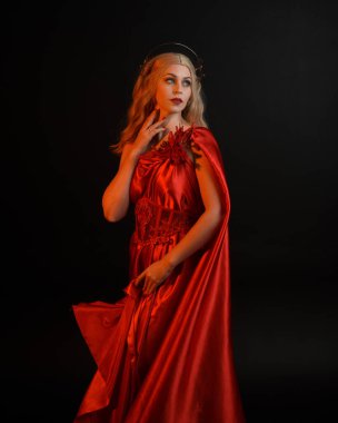 Kırmızı ipek toga elbisesi ve taç giyen, antik mitolojik fantezi tanrıçası gibi giyinmiş güzel sarışın mankenin yakın portresi. Karanlık stüdyo arka planında izole zarif bir poz..