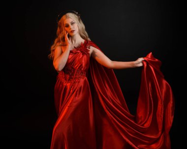 Kırmızı ipek toga elbisesi ve taç giyen, antik mitolojik fantezi tanrıçası gibi giyinmiş güzel sarışın mankenin yakın portresi. Karanlık stüdyo arka planında izole zarif bir poz..