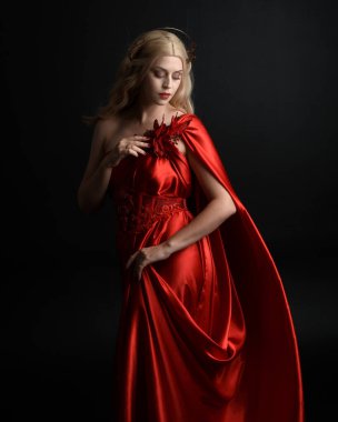 Kırmızı ipek toga elbisesi ve taç giyen, antik mitolojik fantezi tanrıçası gibi giyinmiş güzel sarışın mankenin yakın portresi. Karanlık stüdyo arka planında izole zarif bir poz..