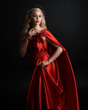 Kırmızı ipek toga elbisesi ve taç giyen, antik mitolojik fantezi tanrıçası gibi giyinmiş güzel sarışın mankenin yakın portresi. Karanlık stüdyo arka planında izole zarif bir poz..
