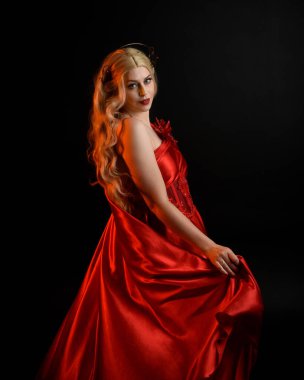 Kırmızı ipek toga elbisesi ve taç giyen, antik mitolojik fantezi tanrıçası gibi giyinmiş güzel sarışın mankenin yakın portresi. Karanlık stüdyo arka planında izole zarif bir poz..
