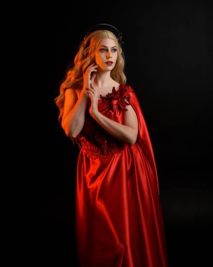 Kırmızı ipek toga elbisesi ve taç giyen, antik mitolojik fantezi tanrıçası gibi giyinmiş güzel sarışın mankenin yakın portresi. Karanlık stüdyo arka planında izole zarif bir poz..