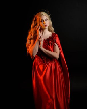 Kırmızı ipek toga elbisesi ve taç giyen, antik mitolojik fantezi tanrıçası gibi giyinmiş güzel sarışın mankenin yakın portresi. Karanlık stüdyo arka planında izole zarif bir poz..