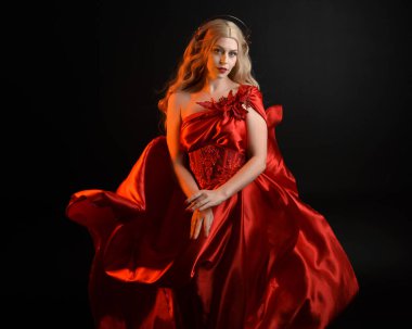 Kırmızı ipek toga elbisesi ve taç giyen, antik mitolojik fantezi tanrıçası gibi giyinmiş güzel sarışın mankenin yakın portresi. Karanlık stüdyo arka planında izole zarif bir poz..