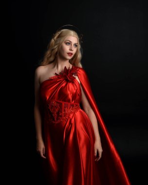Kırmızı ipek toga elbisesi ve taç giyen, antik mitolojik fantezi tanrıçası gibi giyinmiş güzel sarışın mankenin yakın portresi. Karanlık stüdyo arka planında izole zarif bir poz..