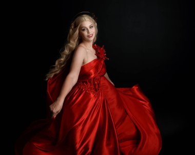 Kırmızı ipek toga elbisesi ve taç giyen, antik mitolojik fantezi tanrıçası gibi giyinmiş güzel sarışın mankenin yakın portresi. Karanlık stüdyo arka planında izole zarif bir poz..