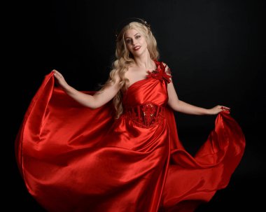 Kırmızı ipek toga elbisesi ve taç giyen, antik mitolojik fantezi tanrıçası gibi giyinmiş güzel sarışın mankenin yakın portresi. Karanlık stüdyo arka planında izole zarif bir poz..