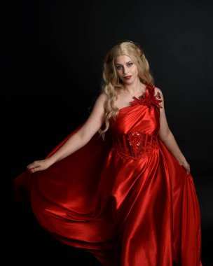 Kırmızı ipek toga elbisesi ve taç giyen, antik mitolojik fantezi tanrıçası gibi giyinmiş güzel sarışın mankenin yakın portresi. Karanlık stüdyo arka planında izole zarif bir poz..
