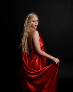 Kırmızı ipek toga elbisesi ve taç giyen, antik mitolojik fantezi tanrıçası gibi giyinmiş güzel sarışın mankenin yakın portresi. Karanlık stüdyo arka planında izole zarif bir poz..