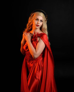 Kırmızı ipek toga elbisesi ve taç giyen, antik mitolojik fantezi tanrıçası gibi giyinmiş güzel sarışın mankenin yakın portresi. Karanlık stüdyo arka planında izole zarif bir poz..