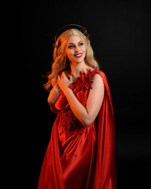 Kırmızı ipek toga elbisesi ve taç giyen, antik mitolojik fantezi tanrıçası gibi giyinmiş güzel sarışın mankenin yakın portresi. Karanlık stüdyo arka planında izole zarif bir poz..