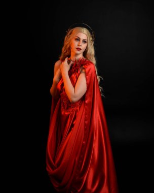 Kırmızı ipek toga elbisesi ve taç giyen, antik mitolojik fantezi tanrıçası gibi giyinmiş güzel sarışın mankenin yakın portresi. Karanlık stüdyo arka planında izole zarif bir poz..