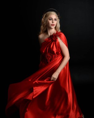 Kırmızı ipek toga elbisesi ve taç giyen, antik mitolojik fantezi tanrıçası gibi giyinmiş güzel sarışın mankenin yakın portresi. Karanlık stüdyo arka planında izole zarif bir poz..
