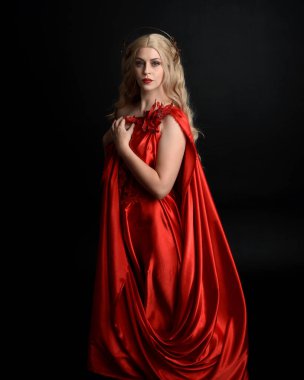 Kırmızı ipek toga elbisesi ve taç giyen, antik mitolojik fantezi tanrıçası gibi giyinmiş güzel sarışın mankenin yakın portresi. Karanlık stüdyo arka planında izole zarif bir poz..