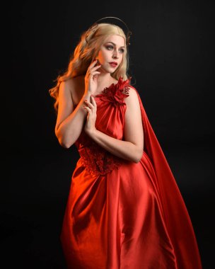 Kırmızı ipek toga elbisesi ve taç giyen, antik mitolojik fantezi tanrıçası gibi giyinmiş güzel sarışın mankenin yakın portresi. Karanlık stüdyo arka planında izole zarif bir poz..