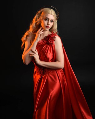 Kırmızı ipek toga elbisesi ve taç giyen, antik mitolojik fantezi tanrıçası gibi giyinmiş güzel sarışın mankenin yakın portresi. Karanlık stüdyo arka planında izole zarif bir poz..