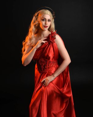 Kırmızı ipek toga elbisesi ve taç giyen, antik mitolojik fantezi tanrıçası gibi giyinmiş güzel sarışın mankenin yakın portresi. Karanlık stüdyo arka planında izole zarif bir poz..