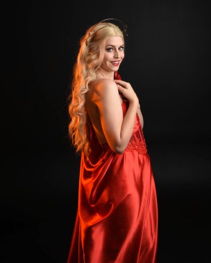 Kırmızı ipek toga elbisesi ve taç giyen, antik mitolojik fantezi tanrıçası gibi giyinmiş güzel sarışın mankenin yakın portresi. Karanlık stüdyo arka planında izole zarif bir poz..