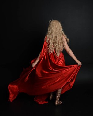 Kırmızı ipek toga elbisesi ve taç giyen, antik mitolojik fantezi tanrıçası gibi giyinmiş güzel sarışın mankenin yakın portresi. Karanlık stüdyo arka planında izole zarif bir poz..