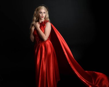 Kırmızı ipek toga elbisesi ve taç giyen, antik mitolojik fantezi tanrıçası gibi giyinmiş güzel sarışın mankenin yakın portresi. Karanlık stüdyo arka planında izole zarif bir poz..