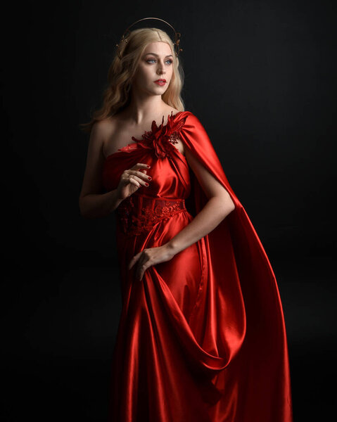 Крупный план портрета красивой блондинки в красном шелковом халате и короне, одетой как древняя мифологическая богиня фантазий. Изящная элегантная поза изолирована на темном фоне студии.